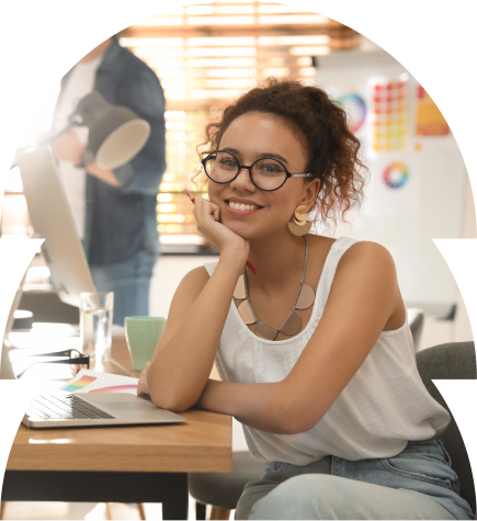 Smiling young woman, looking at the camera, sitting at her desk at work with a laptop in-front of her enjoying do what she is doing